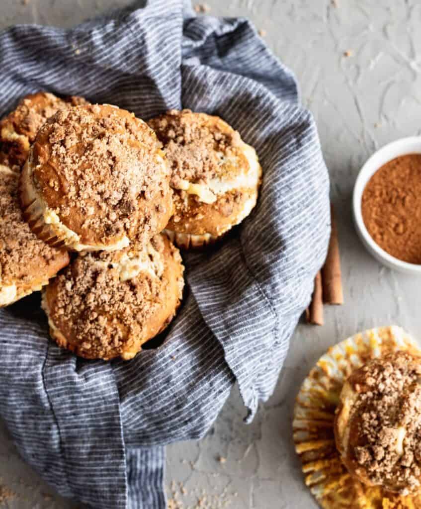 A bowl of pumpkin cheesecake swirl muffins with a cinnamon crumb topping with 1 muffin on the side.