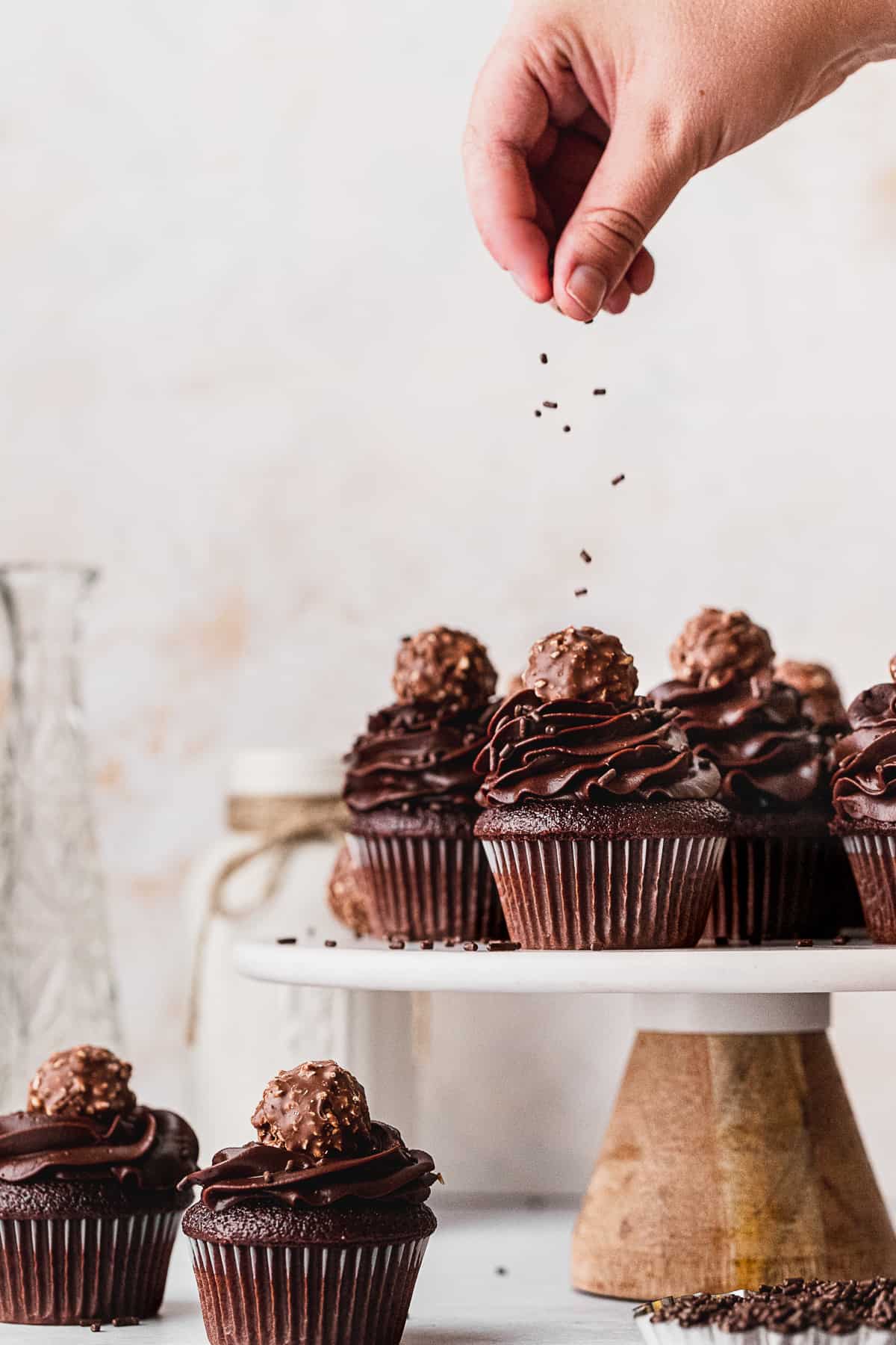 Sprinkling chocolate sprinkles on top of chocolate fudge cupcakes.