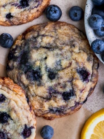 Three lemon blueberry cookies on parchment paper with fresh blueberries around it.