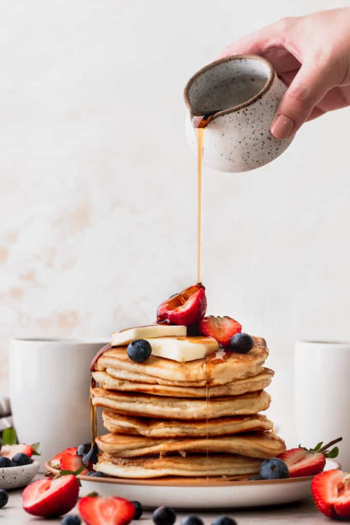 Pouring syrup on top of big stack of pancakes.