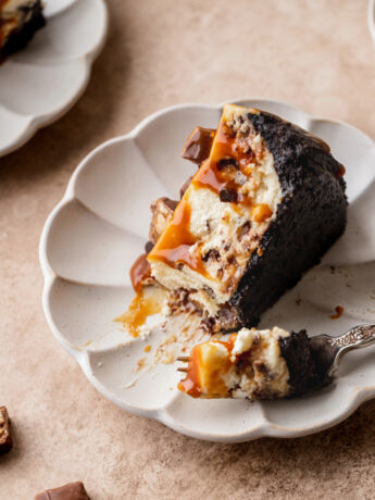 A slice of Snickers cheesecake on its side on a plate.